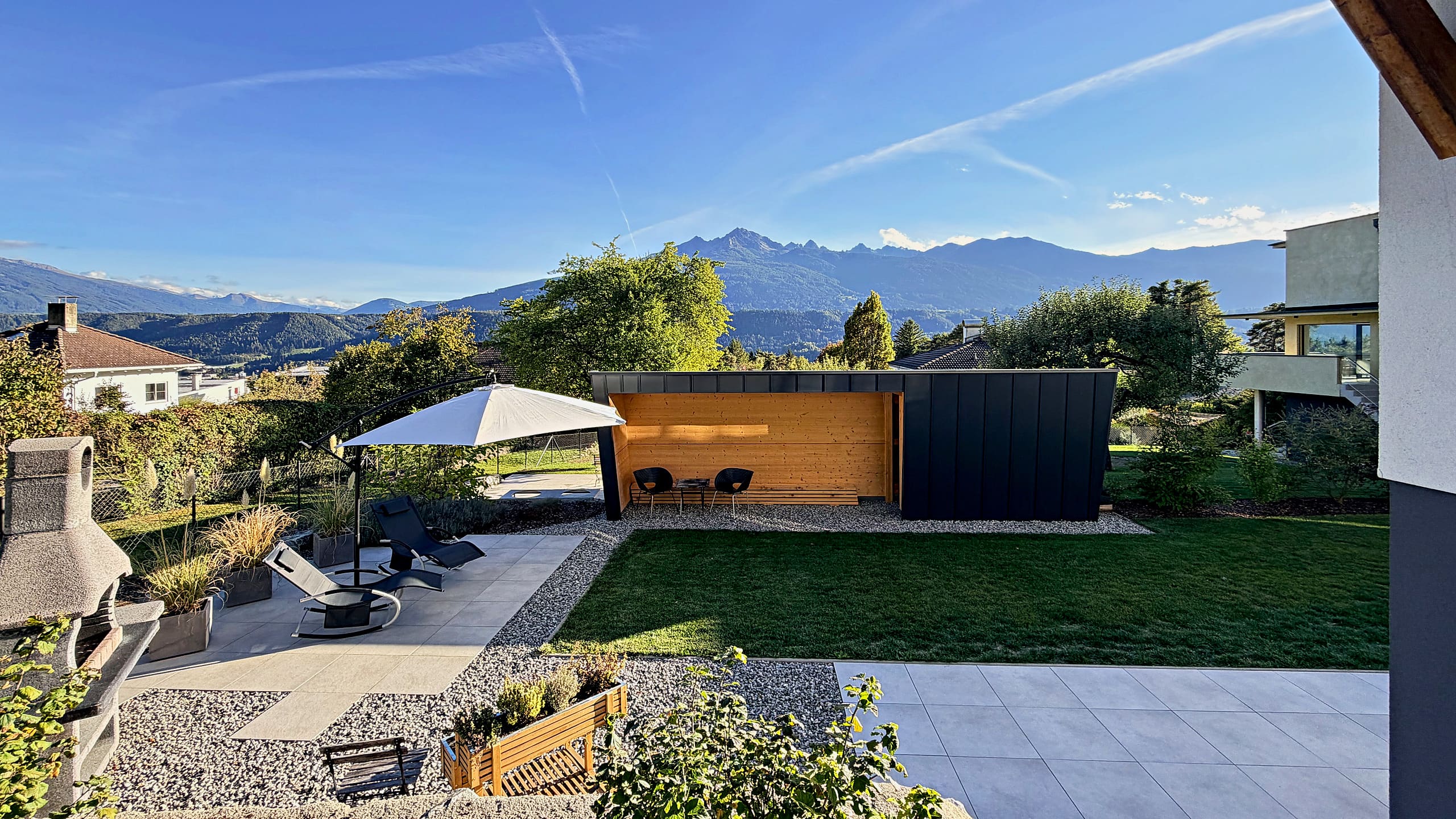 Garten mit Terrasse, Sonnenliegen, Sonnenschirm und gemauertem Grill, dahinter das Bergpanorama über dem Inntal
